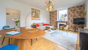 A modern living room with a wooden dining table, a sofa, a TV, and a fireplace. - Le Magda – T2 lumineux entre centre & commerces (Vannes)