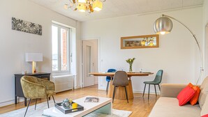 A modern living room with a sofa, coffee table, and chairs. There is a dining table with chairs and a lamp. A painting is on the wall, and a window provides natural light. - Le Magda – T2 lumineux entre centre & commerces (Vannes)