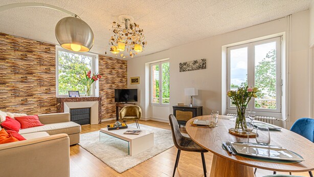 A modern living room with a fireplace, a dining area with a round table, and large windows offering a view of greenery. - Le Magda – T2 lumineux entre centre & commerces (Vannes)