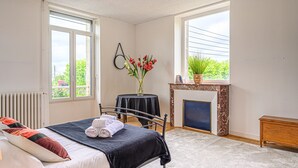A bedroom with a bed, a small table with a vase of flowers, a fireplace, and large windows. - Le Magda – T2 lumineux entre centre & commerces (Vannes)