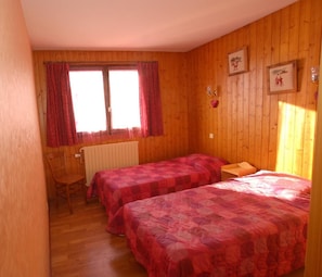 Habitación con paredes de madera y dos camas con ropa de cama roja. Hay una silla y una mesita.