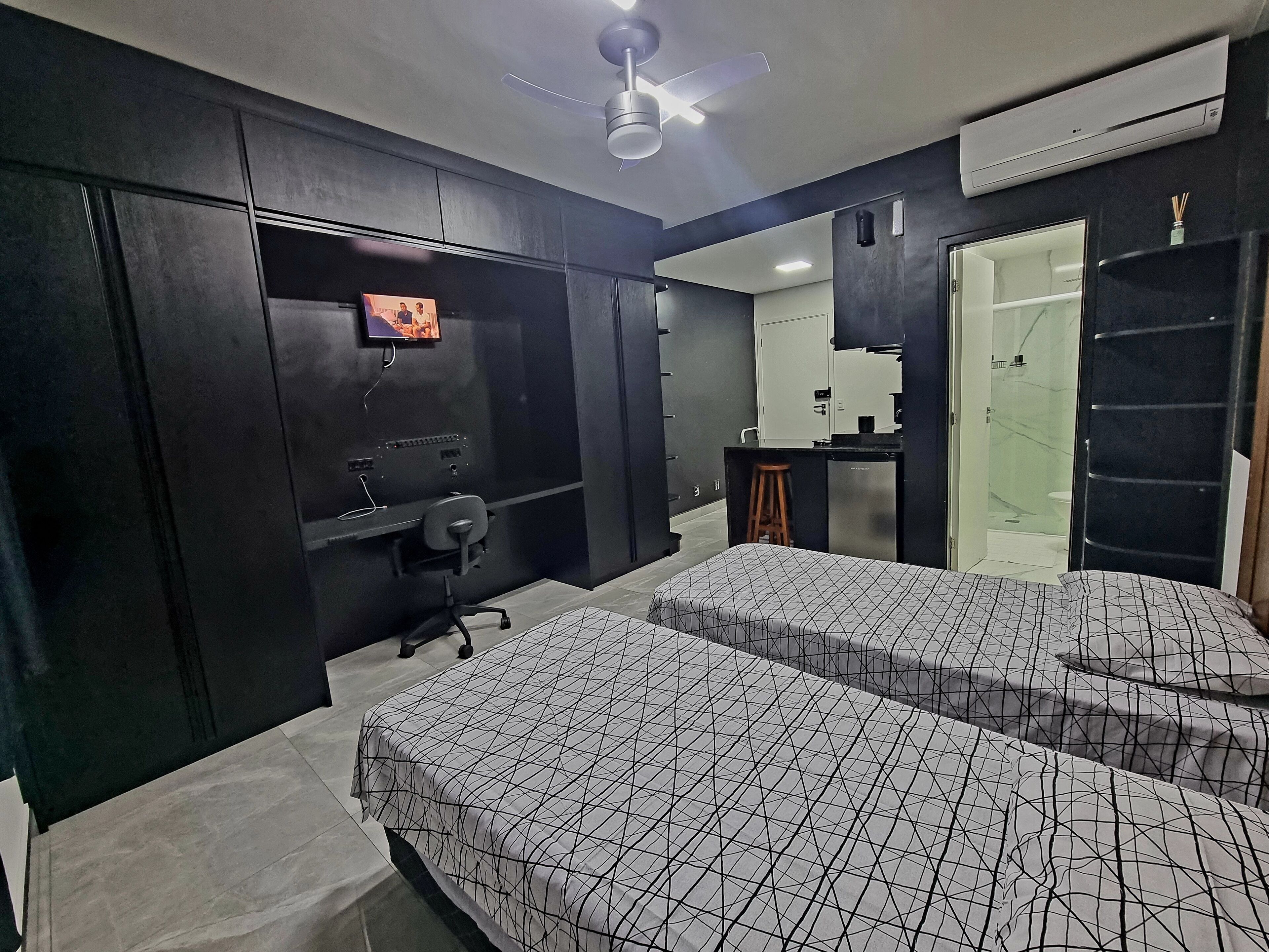 Un dormitorio moderno con dos camas, un escritorio, un televisor y un baño visible a través de una puerta de vidrio.