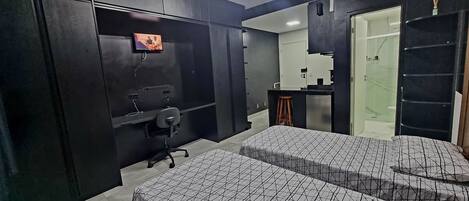Un dormitorio moderno con dos camas, un escritorio, un televisor y un baño visible a través de una puerta de vidrio.