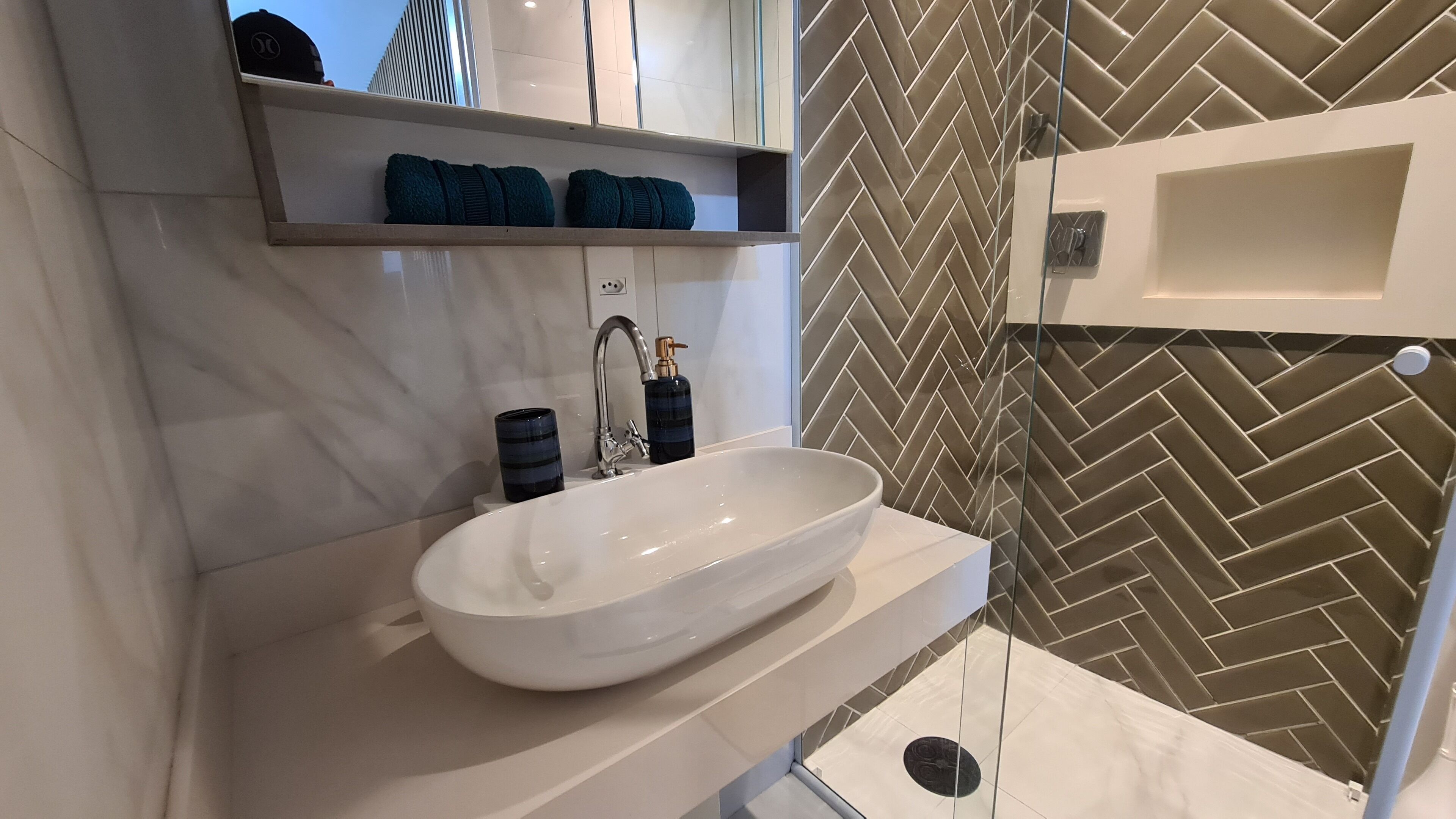 Un baño moderno con un lavamanos blanco, un perchero con toallas dobladas, una cabina de ducha de vidrio y un revestimiento de pared con estampado de espiga.