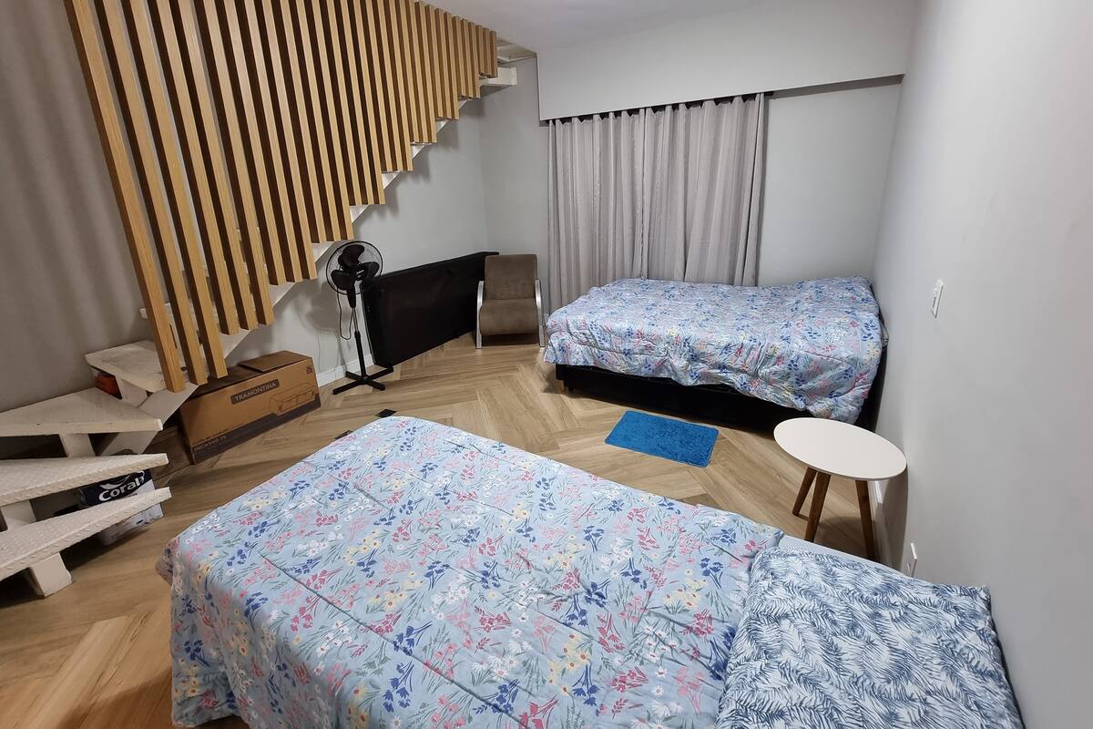 Une pièce avec deux lits, un mur avec des lambris en bois, un ventilateur sur pied, une chaise, une petite table et un tapis bleu.