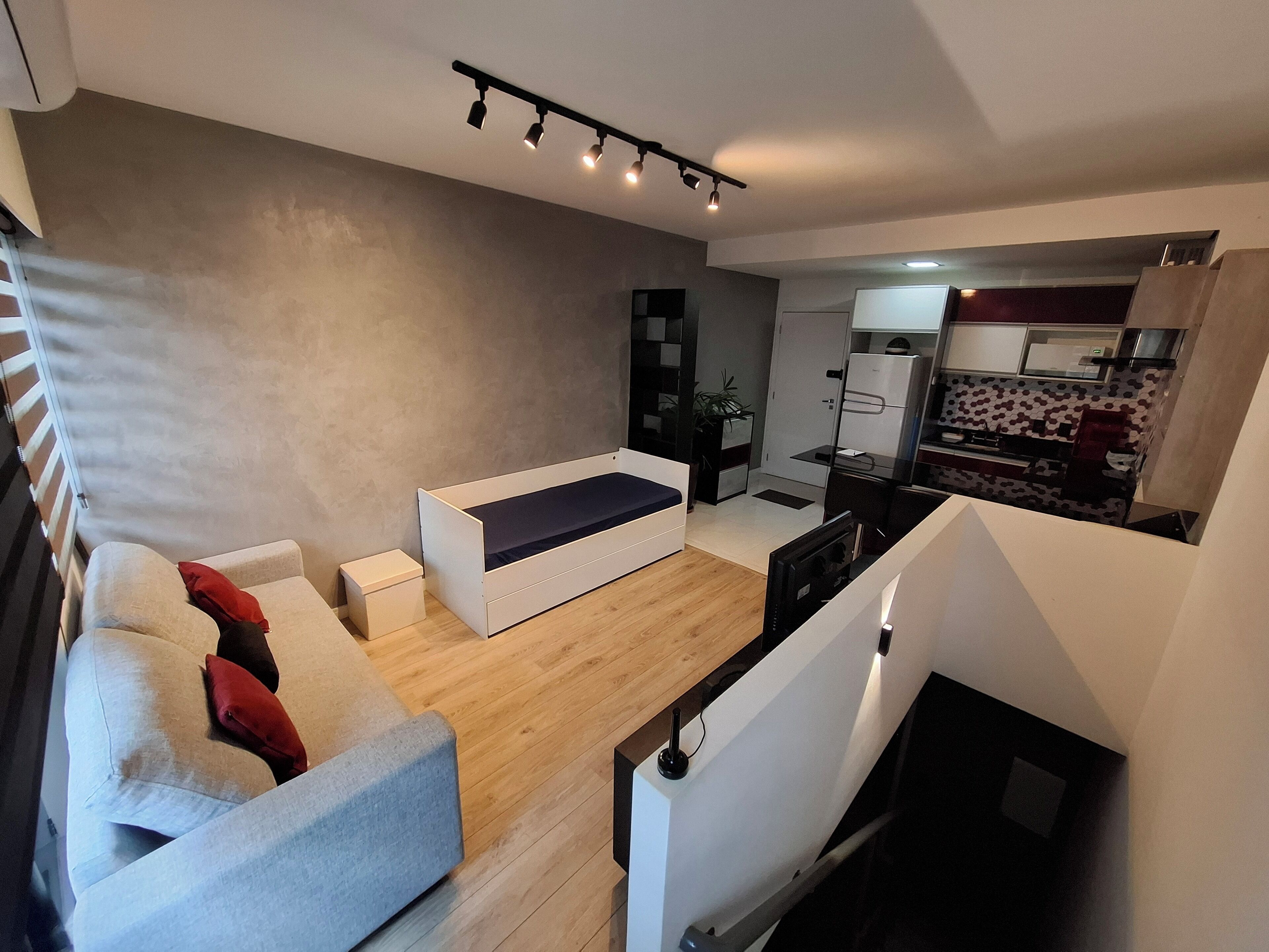 Ein modernes Wohnzimmer mit einer Couch, einem kleinen Tisch und einem Fernseher. Es gibt eine Küchenzeile mit einem Kühlschrank und einer Mikrowelle.