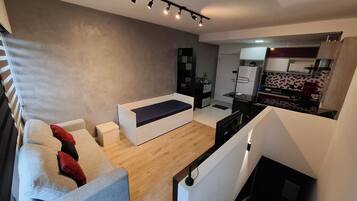 Ein modernes Wohnzimmer mit einer Couch, einem kleinen Tisch und einem Fernseher. Es gibt eine Küchenzeile mit einem Kühlschrank und einer Mikrowelle.