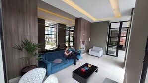 A modern living room with a blue sofa, a white armchair, and a black coffee table. There is a potted plant and a mirror on a small table. - Edificio Selenita Loft e Duplex (Barueri)