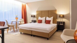 A hotel room with a large bed, two bedside tables with lamps, a small round table with chairs, and a view of the outdoors through sheer curtains. - Livingroom Mathildenhöhe (Darmstadt)