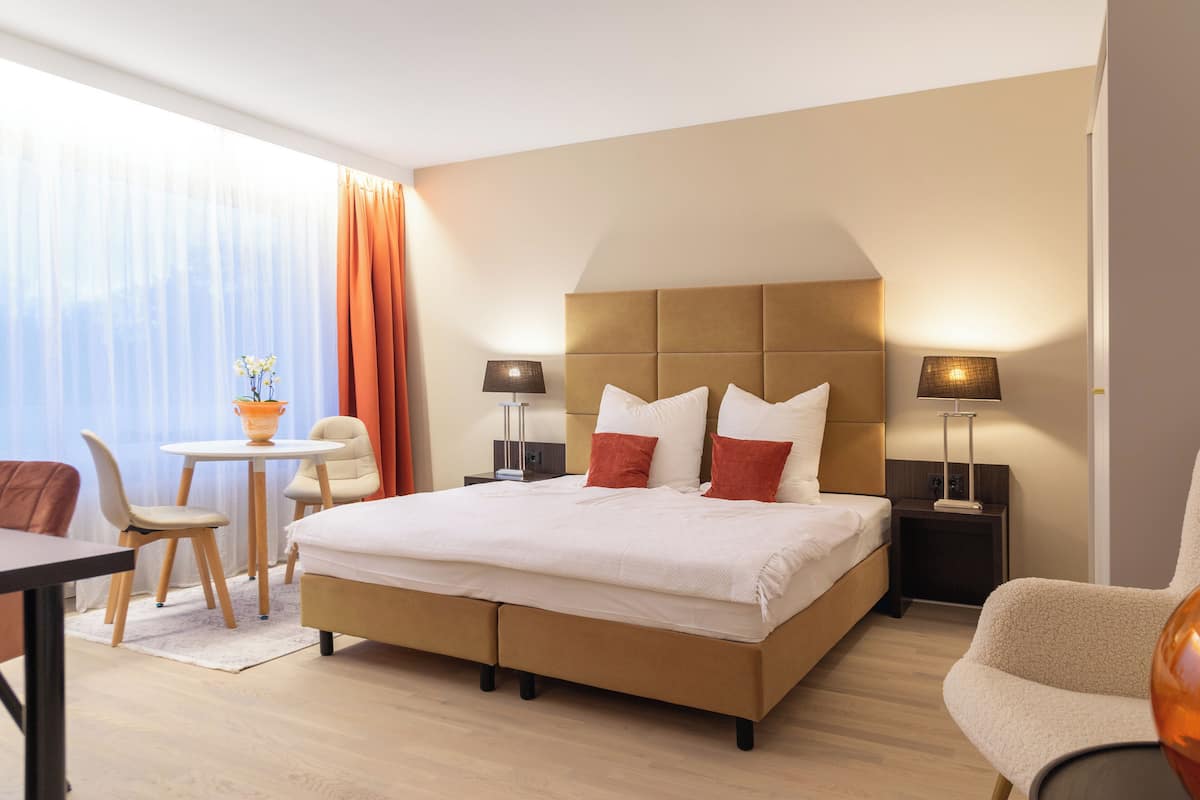 A hotel room with a large bed, two bedside tables with lamps, a small round table with chairs, and a view of the outdoors through sheer curtains.