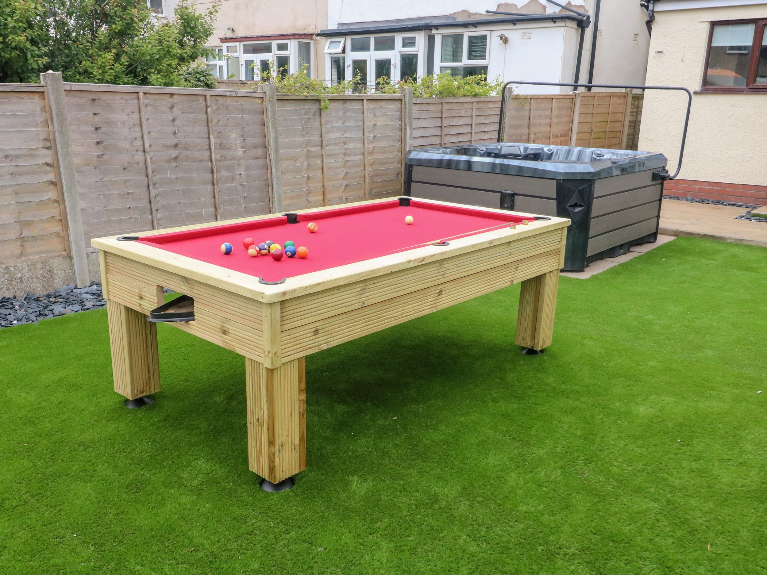 A pool table with balls on a green lawn, surrounded by a wooden fence and a hot tub.