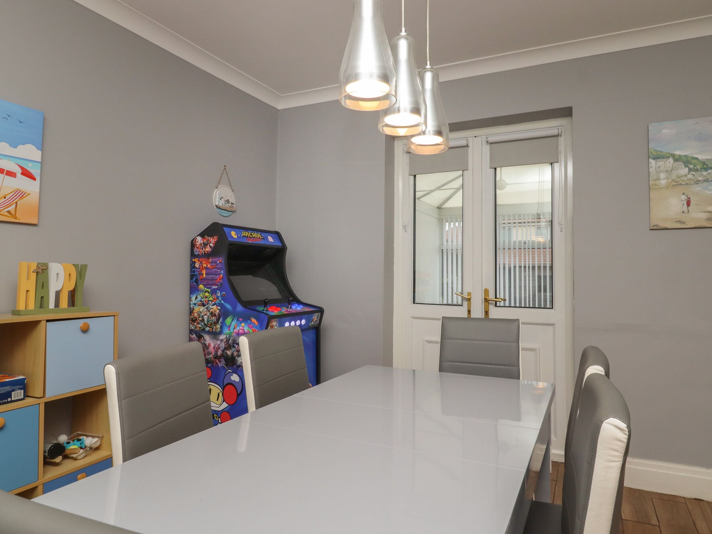 A dining area with a white table, grey chairs, a colorful arcade machine, and a view through a door.