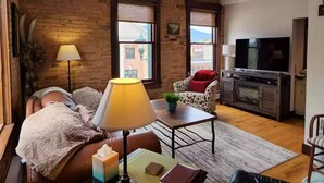 A cozy living room with a brick wall, a sofa, a coffee table, a TV, and a fireplace. - Charming and Beautiful Downtown Whitefish Condo in Historic Building (Whitefish)