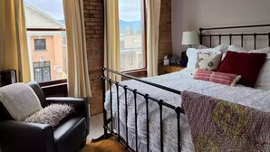 A bedroom with a bed, a chair, a nightstand, and a window with curtains. - Charming and Beautiful Downtown Whitefish Condo in Historic Building (Whitefish)