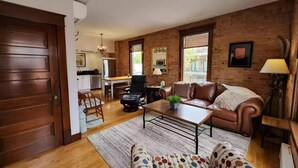 A cozy living room with a wooden floor, a leather sofa, a patterned armchair, and a dining area with a table and chairs. - Charming and Beautiful Downtown Whitefish Condo in Historic Building (Whitefish)