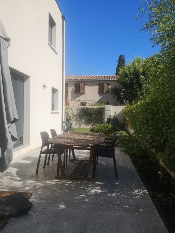 Un patio con una mesa y sillas redondas, un edificio blanco con techo de tejas y un jardín con diversas plantas.