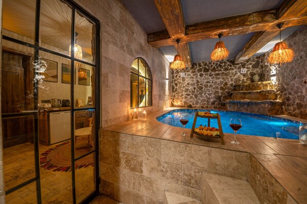 A stone-walled indoor pool area with wooden beams, a glass partition, and a fruit bowl with wine glasses. - Louis Cappadocia (Nevsehir)