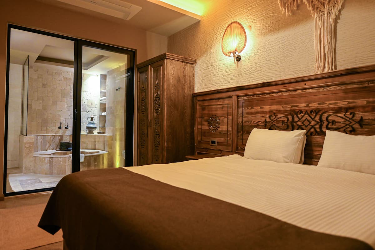 A hotel room with a large bed, a wooden headboard, a wall-mounted light fixture, and a sliding glass door leading to a bathroom.