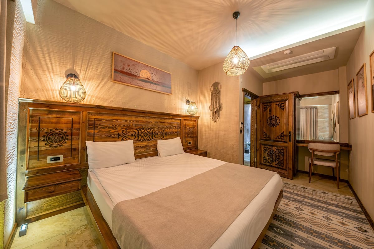 A well-lit bedroom with a large bed, wooden headboard, bedside tables, a desk, and a chair.