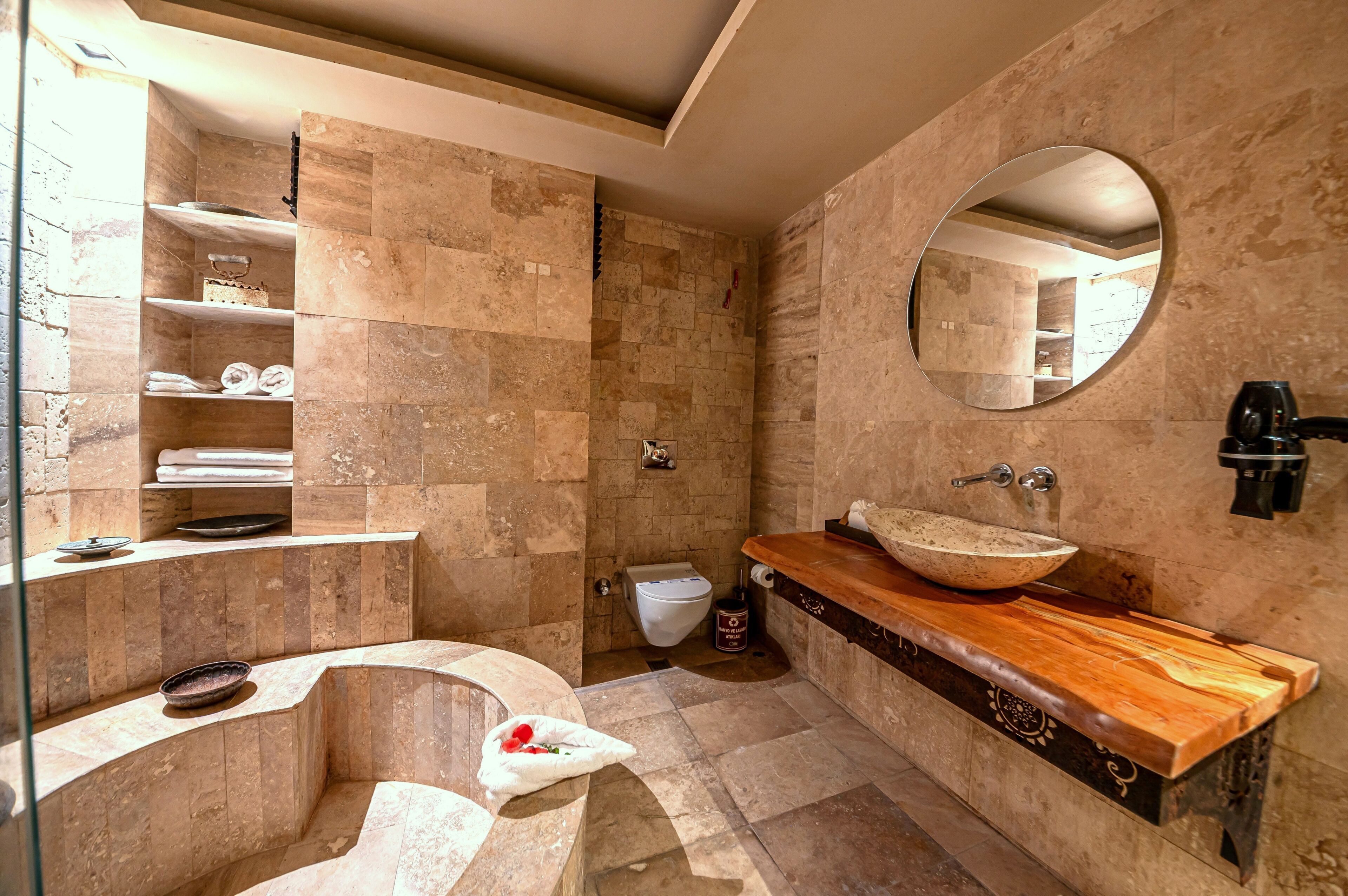 A bathroom with a stone tile wall, a wooden vanity, a round mirror, and a built-in shower.