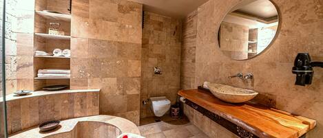 A bathroom with a stone tile wall, a wooden vanity, a round mirror, and a built-in shower.