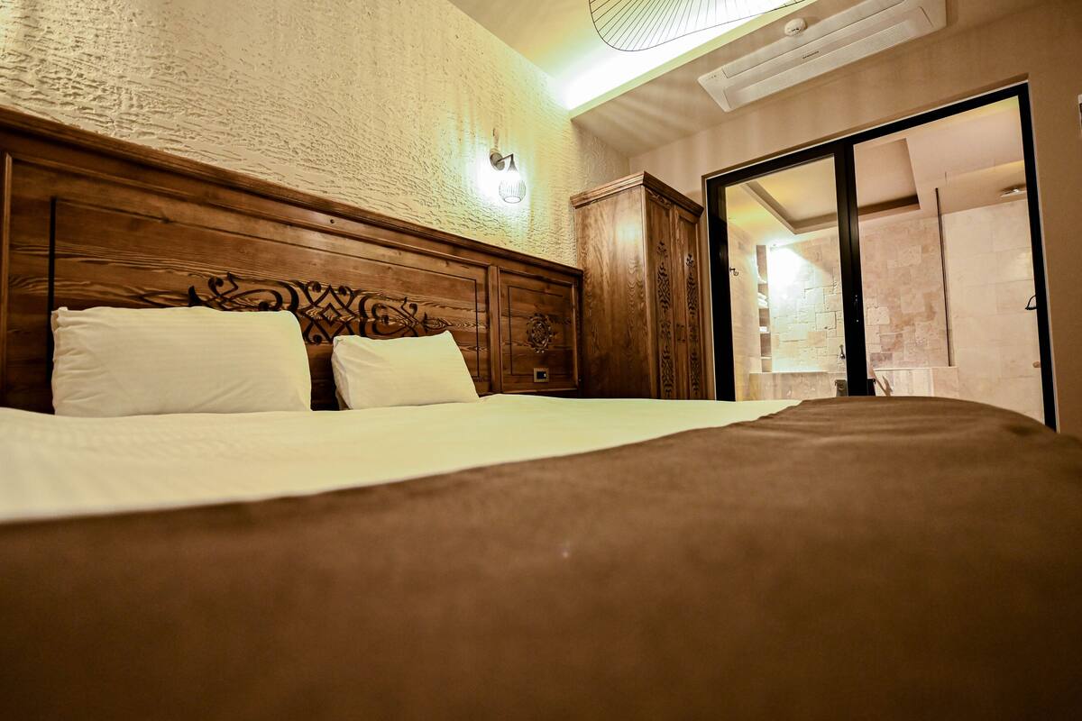 A bedroom with a wooden headboard, a bed with white linens, a wardrobe, and a sliding glass door leading to a bathroom.
