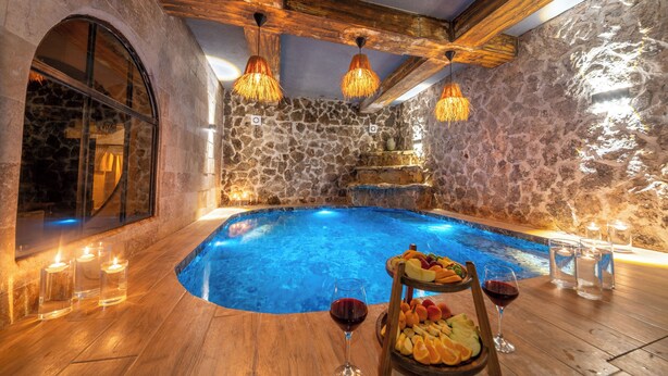 A stone-walled indoor pool with wooden beams, candles, and fruit on a table.