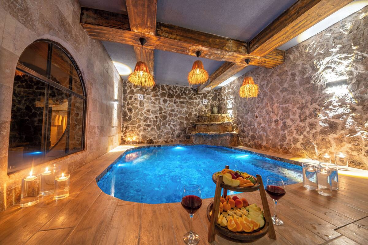 A stone-walled indoor pool with wooden beams, candles, and fruit on a table.