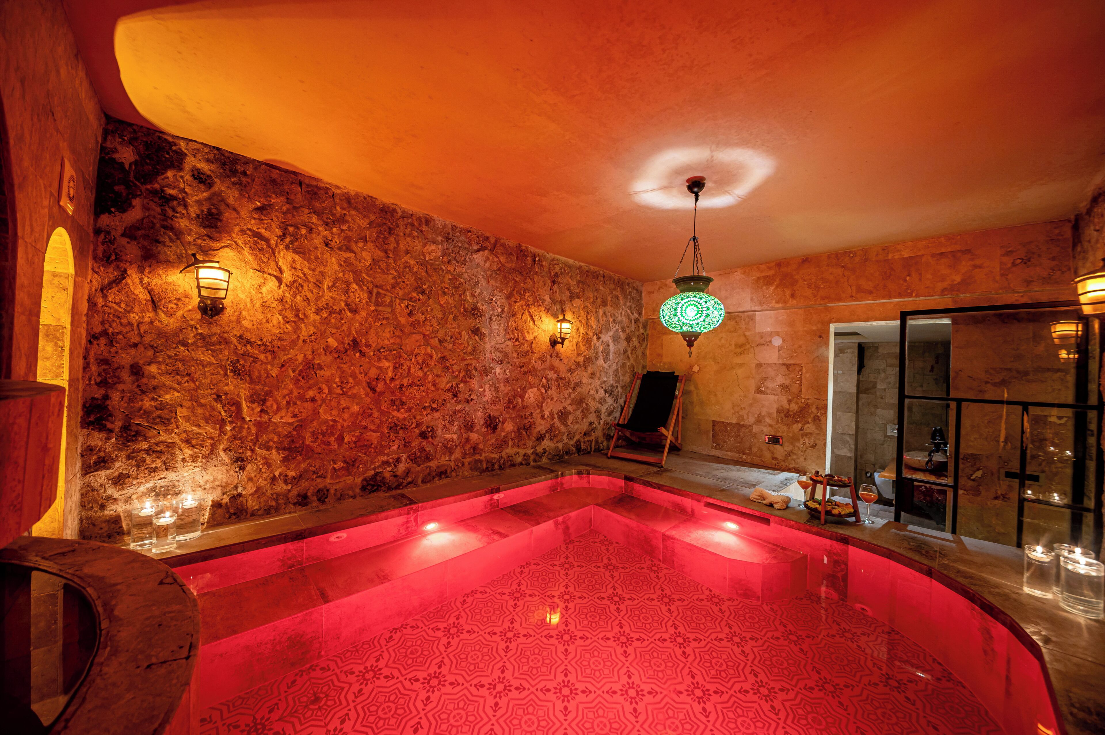 Una habitación con paredes de piedra, una piscina con luz roja, un barril de madera y una puerta de cristal que da a otra zona.