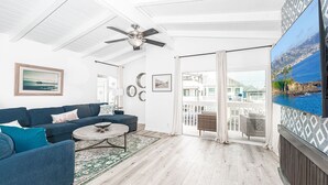 A spacious living room with a blue sofa, a coffee table, and a ceiling fan. There is a large window with a view of buildings and a balcony. - Remodeled & Stylish Upper-Level Home, Steps to the Sand! (Newport Beach)