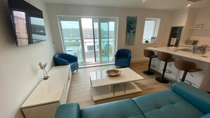 A modern living room with a blue sofa, a white coffee table, and a blue armchair. There is a kitchen area with a bar and stools. - Stunning Cliff Edge Apartment in Newquay (Newquay)