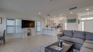 A spacious living room with a grey sofa, a coffee table, and a TV. There is a kitchen area with white cabinets and a dining area with white chairs.