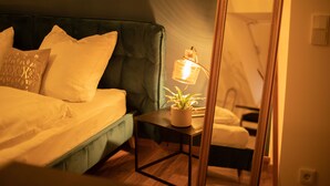 Un dormitorio con cabecera tapizada de verde, una mesita de noche con lámpara y una planta en maceta.