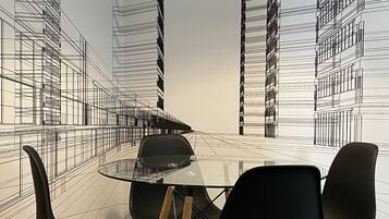 A dining area with a glass table surrounded by chairs, a hanging pendant light, and a wall with a black and white architectural line drawing.