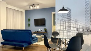 Ein modernes Wohnzimmer mit einem blauen Sofa, einem Esstisch mit Stühlen, einem an der Wand montierten Fernseher und einem großen Fenster mit Blick auf die Stadt.