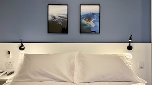A bed with white linens, a bedside table with a phone and remote, and two framed pictures on the wall. - ADB Royal Brooklin By Anora Spaces (São Paulo)