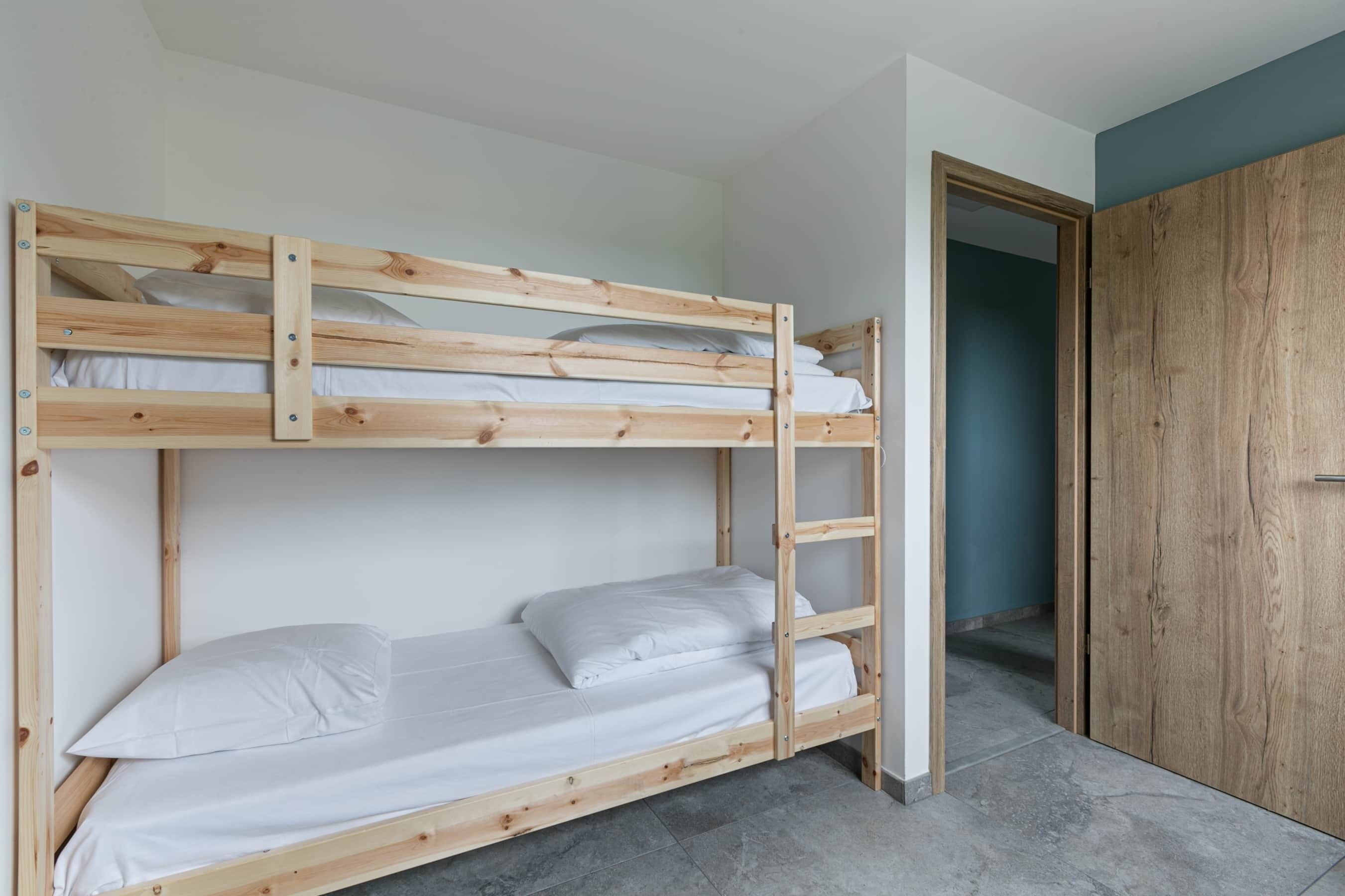 Een stapelbed met witte beddengoed, een houten trap en een garderobe in de aangrenzende kamer.
