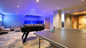 Un espacio interior moderno con mesa de ping pong, mesa de fútbol y una zona de estar con sofá y una mesita.