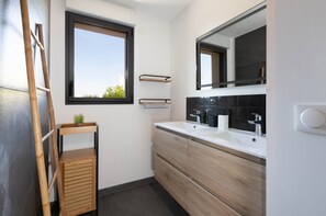 A modern bathroom with a large window, wooden shelves, a mirror, and a sink with black tiles.