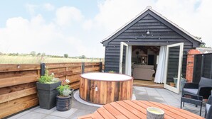 Une terrasse en bois avec une table ronde et des chaises en bois, une petite structure en bois dotée d’une porte en verre, et des plantes en pot.