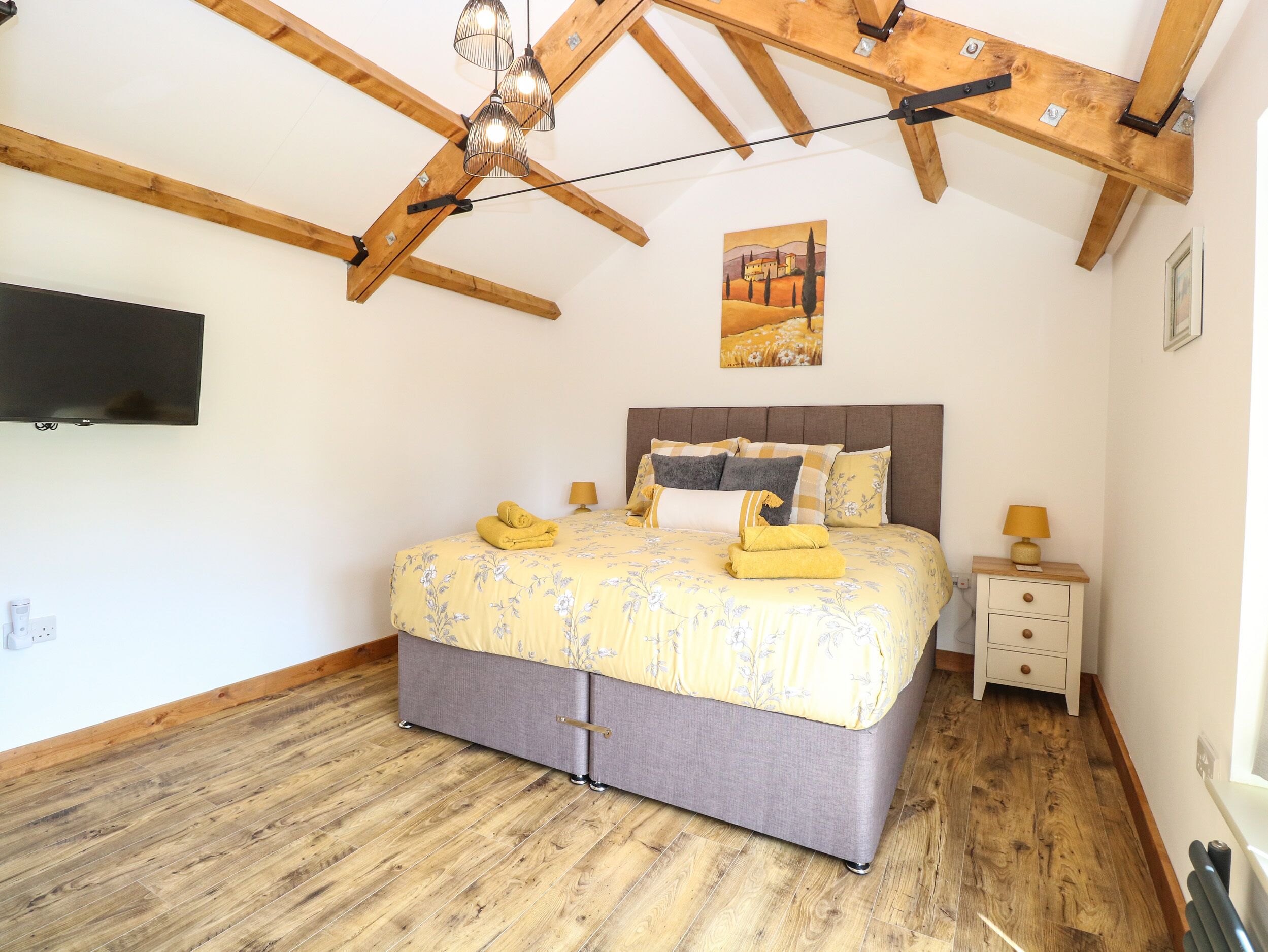 A bedroom with a bed, bedside tables, a TV, wooden beams, and a painting on the wall.