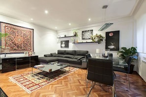 A modern living room with a sofa, coffee table, and a glass dining table. There are plants, artwork, and a patterned rug on the floor.