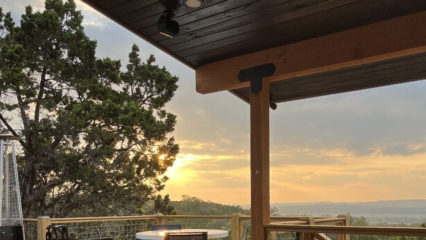 Una terraza de madera con un sofá, un reposapiés, una mesa redonda y vistas al atardecer sobre un paisaje montañoso.