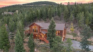 A large wooden house surrounded by dense forest, with a prominent deck and multiple windows.