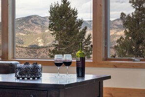 A table with two wine glasses and a bottle of wine, set against a backdrop of mountains and trees through a window.