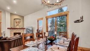 A dining area with a wooden table set for six, a fireplace, and a view of trees through large windows.