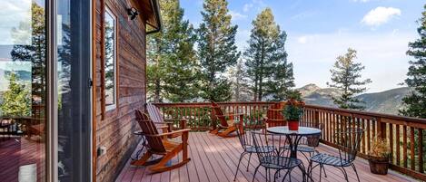 A wooden deck with outdoor furniture, including rocking chairs and a table, overlooking a mountainous landscape.