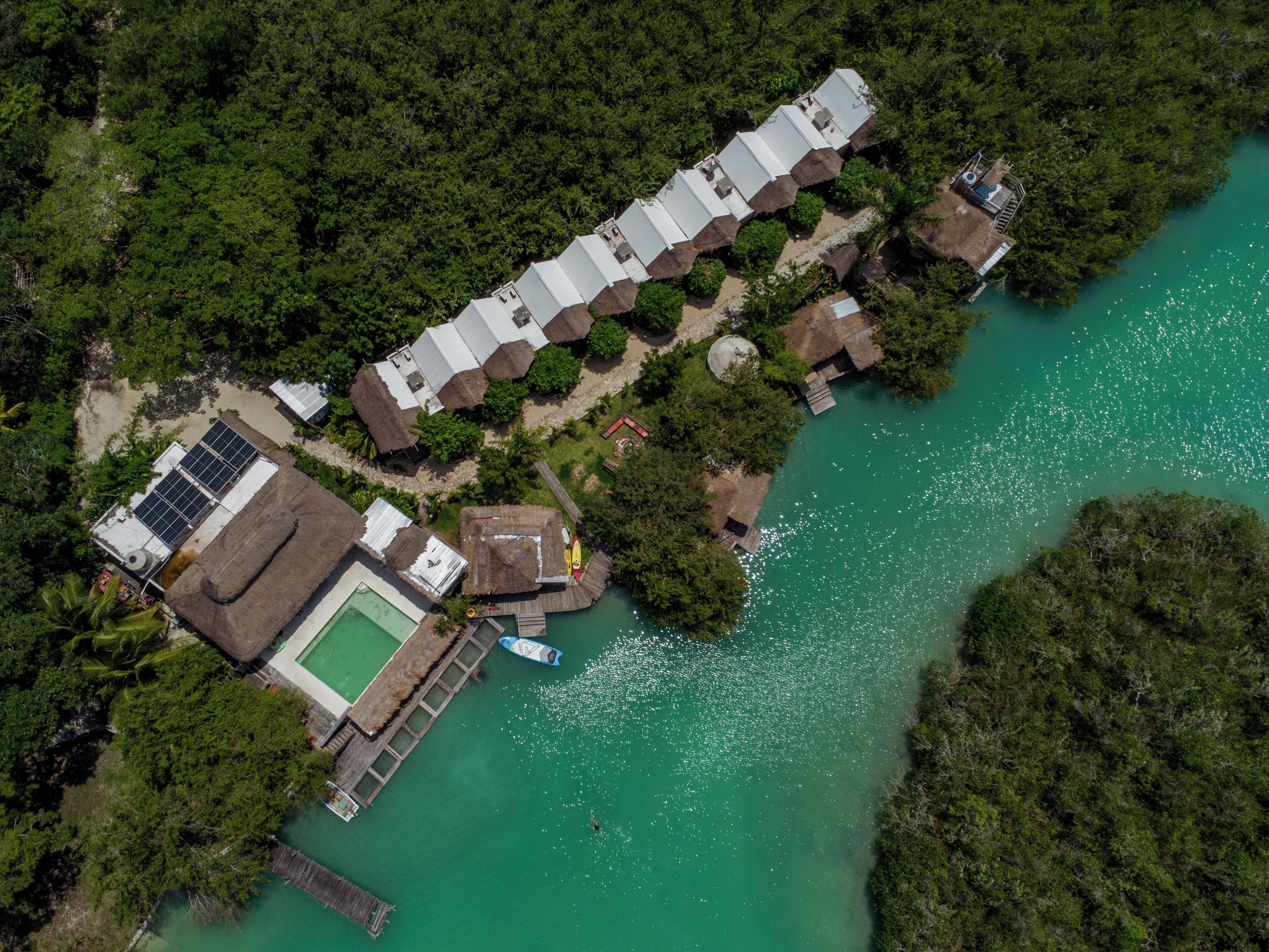 An aerial view of a resort with a long building, a swimming pool, and a dock extending into a turquoise body of water.