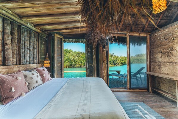 A wooden room with a bed, a hanging light fixture, and a view of a body of water. - Embajada Universal (Huay-Pix)