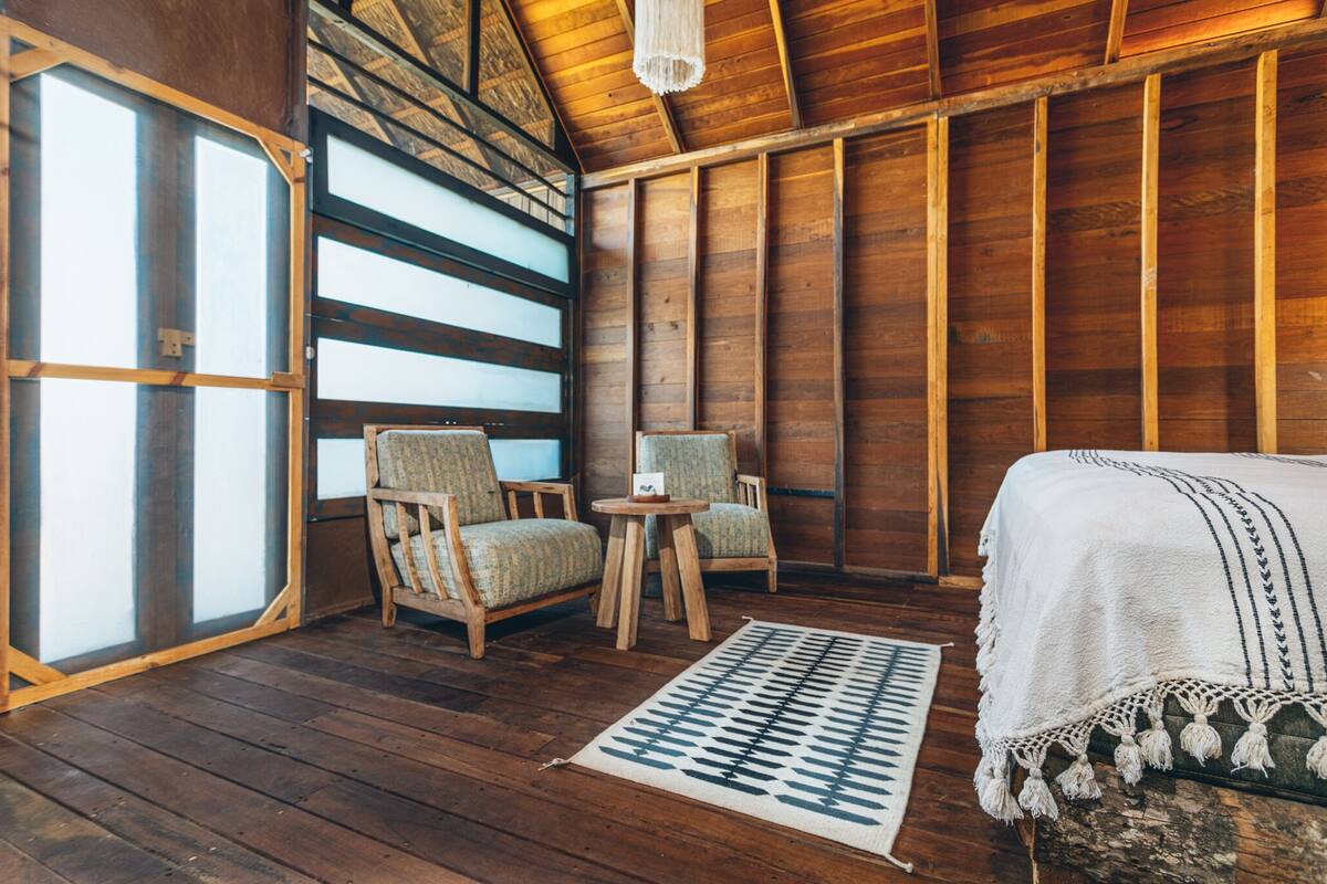 Ein Zimmer aus Holz mit einem Bett, zwei Stühlen, einem kleinen Tisch und einem großen Fenster.